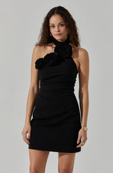 A woman is wearing a sleeveless black dress with a high neckline. The dress features large fabric roses on the left shoulder, adding a decorative element. She is accessorized with gold bracelets on her right wrist and rings on both hands.